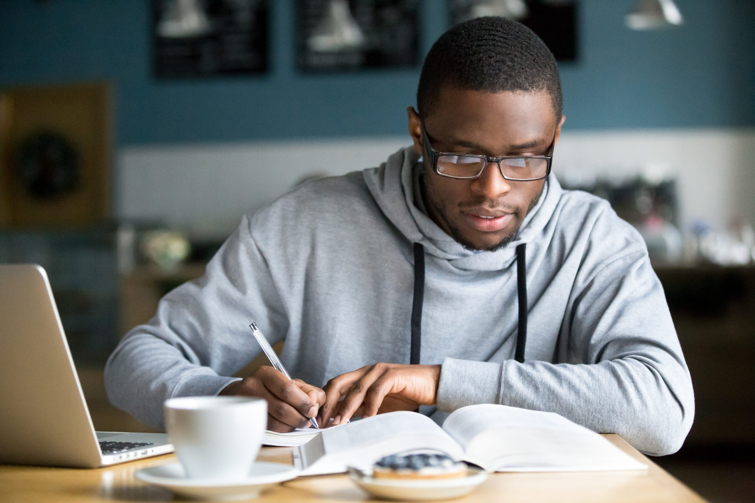 Un homme qui prend des notes sur un livre avec un ordinateur devant lui et une tasse.