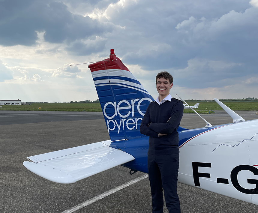 Un jeune homme posant souriant au premier plan, devant un avion portant le logo d'Aéropyrénées.